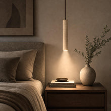 Slender natural travertine pendant light with downward glow, walnut cord, above a bedside table.
