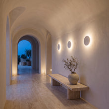 A modern interior hallway with arched ceilings and walls, warm wall-mounted circular lights, a light wood bench, and a ceramic vase with dried branches. The scene appears to be a real architectural environment rather than a clean isolated product shot.