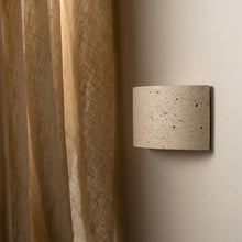Compact Pulse Travertine Up/Down Wall Light, a textured beige stone fixture casting light on a neutral wall next to a linen curtain.