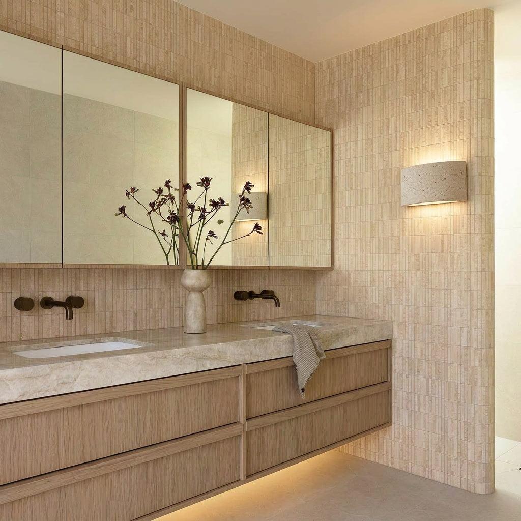 Warm travertine Pulse Up/Down Wall Light illuminating a modern bathroom with wood vanity and stone tiles.